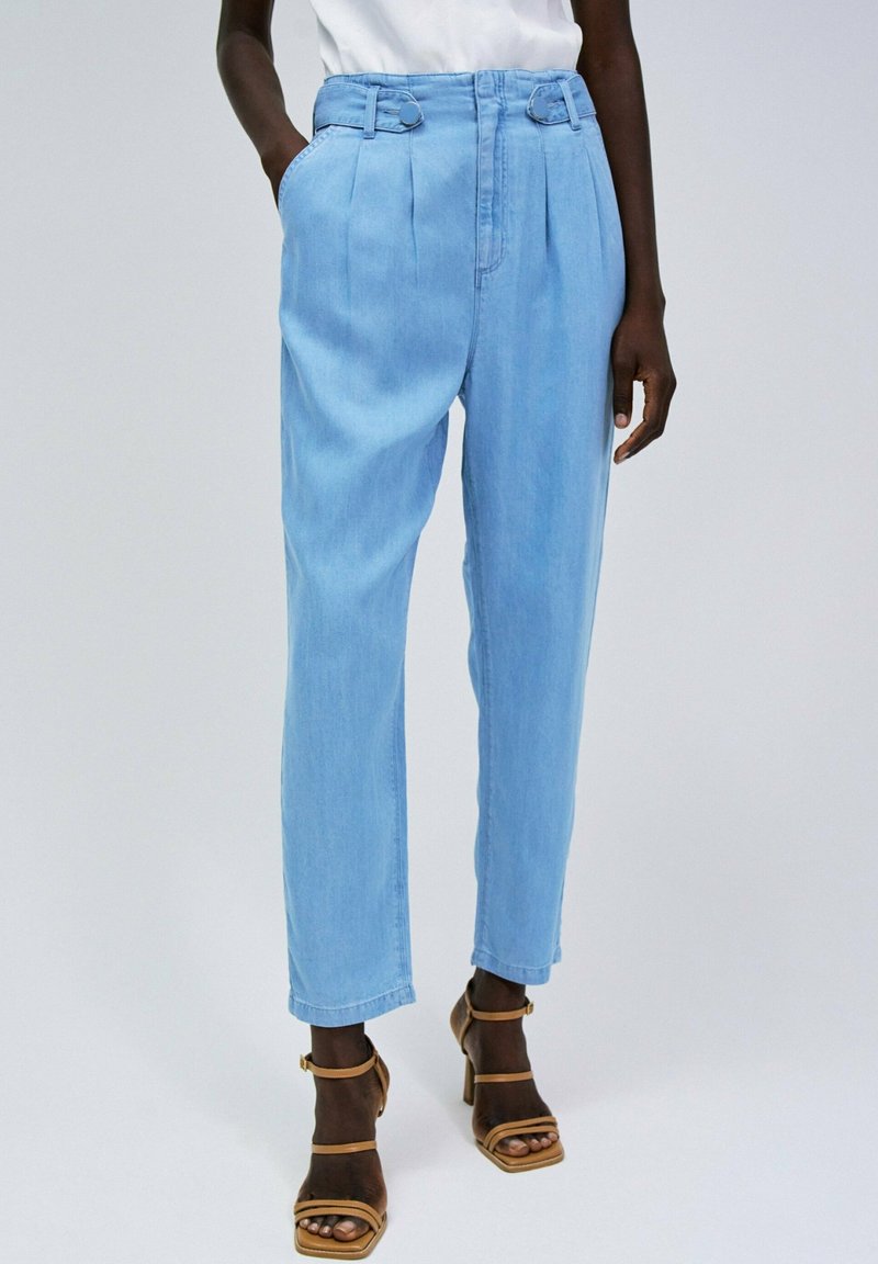 Pantalones de tiro alto azul claro con pliegues y bolsillos laterales, que cuentan con un cinturón con acusaciones de hardware en forma de círculo, combinados con sandalias de tacón marrones.