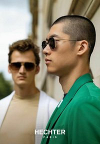 Twee personen met zonnebrillen staan buiten. De een draagt een groene jas, de ander een witte cardigan over een beige shirt.