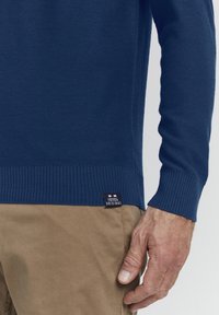 Granatowy sweter z dzianiny, z ribbowymi mankietami i dolnym wykończeniem. Zawiera małą metkę z tkaniny z białym tekstem i wzorem w paski.