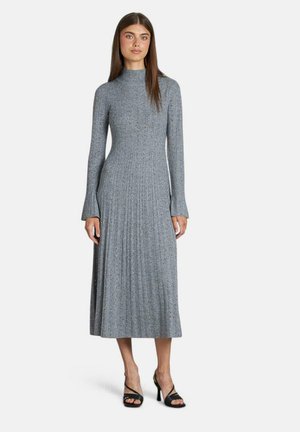 Robe tricotée gris à côtes avec des manches longues et un col montant ; détail de jupe plissée. Le modèle l'associe à des talons noirs à lanières.