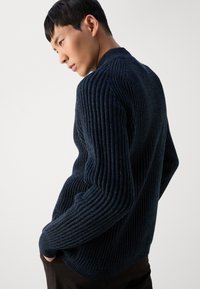 Maglione a coste di un blu navy scuro, realizzato in tessuto lavorato a maglia, con un design aderente e scollatura rotonda, mostrato da un'angolazione laterale.