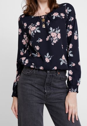 Femme portant un chemisier bleu marine à fleurs manches longues, rentré dans un jean noir taille haute, accessoirisée avec des colliers dorés superposés.