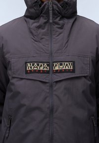 Grå vandtæt jakke med høj krave, lynlås-lukning og to sidelommer med lynlås. Viser et logo-patch på brystområdet.