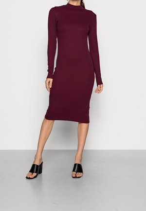 Femme portant une robe midi bordeaux ajustée à manches longues et des sandales noires à talons carrés ouverts, debout devant un fond uni.