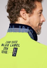 Neon gele jas met een marineblauwe kraag. Bevat geborduurde tekst: "Camp David Blue Label Crew Voyage" en een logo badge.