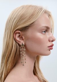 Goldene Metalldesign-Ohrringe mit einem sternförmigen Stecker und mehreren hängenden Sternanhängern, die in einem strukturierten Finish und reflektierenden Oberflächen kreisförmig angeordnet sind.
