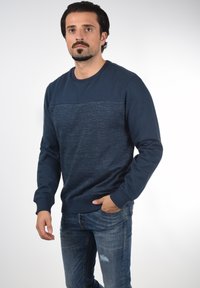 Dunkelblaues Sweatshirt mit strukturierten horizontalen Streifen, Rundhalsausschnitt und gerippten Bündchen. Kombiniert mit verwaschenen blauen Jeans.