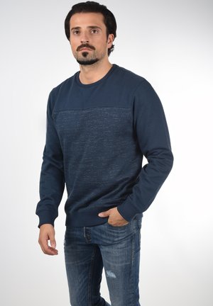 Dunkelblaues Sweatshirt mit strukturierten horizontalen Streifen, Rundhalsausschnitt und gerippten Bündchen. Kombiniert mit verwaschenen blauen Jeans.