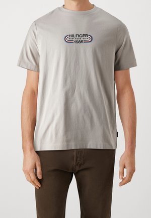 Mężczyzna noszący jasnoszarą koszulkę z krótkim rękawem z logo „Hilfiger New York City 1985” i ciemnobrązowe spodnie, stojący z luźno opuszczonymi ramionami.