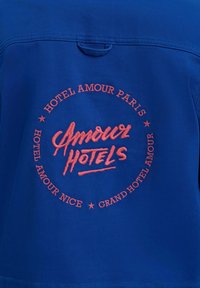 Modro blago z rdečim vezenim napisom "Amour Hotels" na sredini, obdano z napisi "Hotel Amour Paris," "Hotel Amour Nice" in "Grand Hotel Amour."