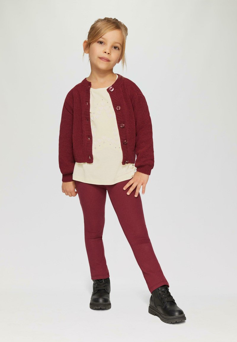 Burgunderfarbener Strickcardigan über einem hellen Creme-Shirt, kombiniert mit taillierten burgunderfarbenen Rippleggings und schwarzen Schnürankelstiefeln.