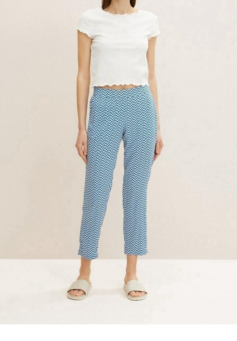 Femme portant un crop top blanc à manches courtes, un pantalon bleu à motifs et des sandales beiges, debout devant un fond uni.