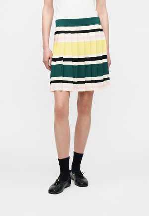 Πλισέ φούστα με οριζόντιες ρίγες σε πράσινο, κίτρινο, μαύρο και απαλό ροζ, φορεμένη με μαύρες κάλτσες και μαύρα μπαλαρινάκια.