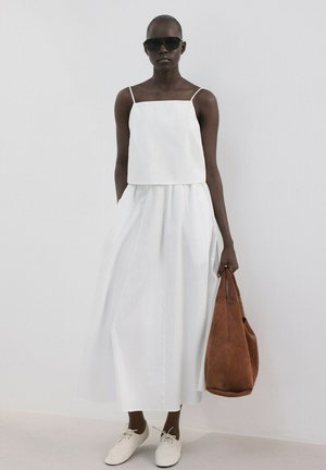 Kobieta ubrana w biały bezrękawnik i długą spódnicę, białe buty, czarne okulary przeciwsłoneczne, trzymająca dużą brązową torbę na białym tle.
