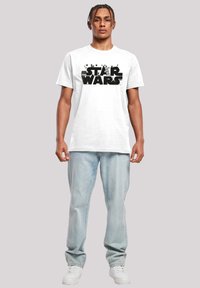 Hvit bomulls-T-skjorte med en dristig svart "STAR WARS" grafikk. Modellen har på seg lyseblå jeans og hvite joggesko. Enkel design, kortermet.