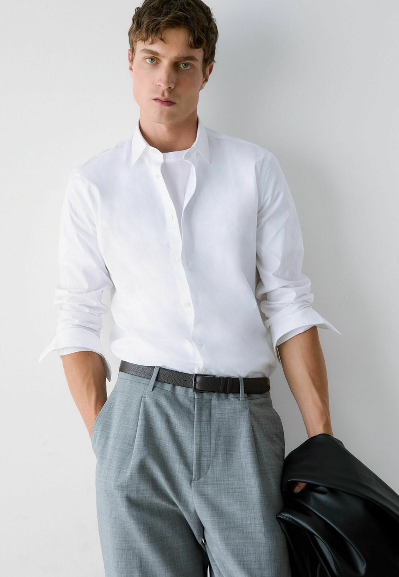 Jeune homme portant une chemise blanche aux manches retroussées, pantalon gris avec ceinture noire, tenant une veste noire dans sa main gauche.