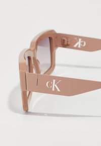 Veľké, svetloružové slnečné okuliare s gradientnými šošovkami a mierne obdĺžnikovým tvarom. Obsahujú logo "CK" na straniciach.