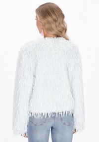 Frau mit blonden, welligen Haaren, die eine hellblaue, fransige, strukturierte Langarmjacke und hellblaue Jeans trägt, von hinten auf weißem Hintergrund.