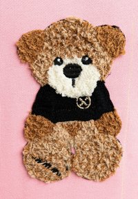 Aplicación de oso de peluche, hecho de tela texturizada marrón y beige con una camiseta negra. Presenta detalles del rostro bordados sobre un fondo rosa.