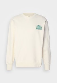 Felpa crema con collo rotondo, maniche lunghe e polsini a coste. Presenta un design grafico teal sul lato sinistro del petto. Tessuto morbido.