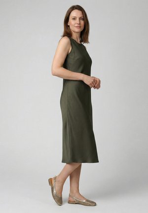 Vrouw staand in zijaanzicht, gekleed in een mouwloze olijfgroene midi-jurk en gouden open achterkant platte schoenen tegen een effen grijze achtergrond.