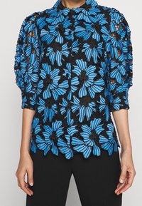 Black blouse featuring blue floral embroidery, with a textured open lace design and a collared neckline. Long sleeves with ruffled accents.
