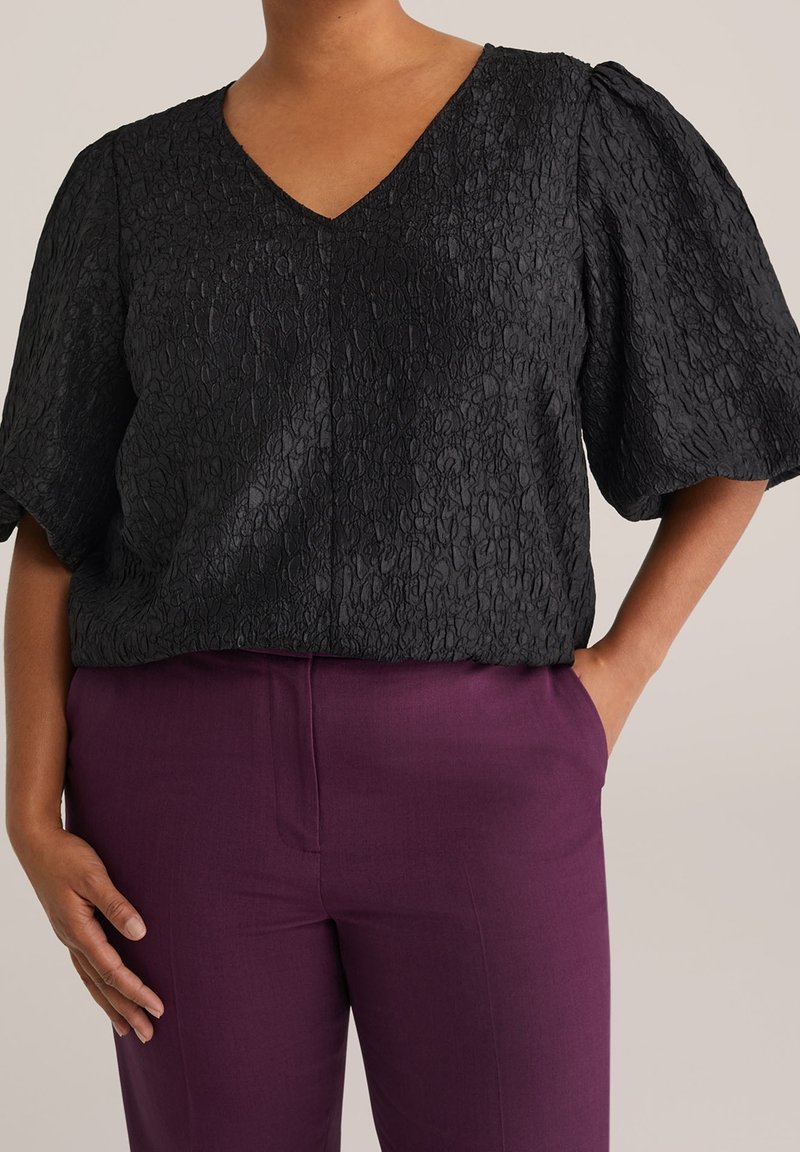 Femme portant un chemisier noir texturé à col en V avec des manches bouffantes et un pantalon violet taille haute, debout avec une main dans la poche.