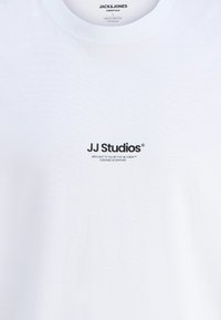 Bijela pamučna majica s malim crnim logotipom "JJ Studios" na prsima. Mekan materijal, dizajn s okruglim izrezom, a označena je kao organski pamuk.