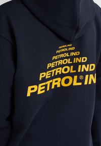 Námořnická mikina s velkým, opakujícím se žlutým logem „PETROL IND“ v různých velikostech, vybavená stahovací kapucí a měkkou texturou.