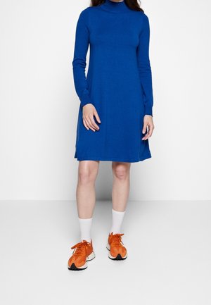 Nainen, joka pukeutunut polvipituiseen kuninkaansiniseen pitkähihaiseen mekkoon, valkoisiin sukkiin ja kirkkaanoransseihin lenkkareihin, seisoo yksinkertaisen taustan edessä.