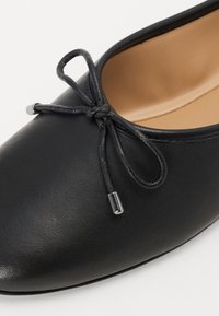 Chaussure plate en cuir noir avec un bout rond, ornée d'un nœud décoratif noué avec de fines lacets et des accents en métal argenté. Texture lisse.