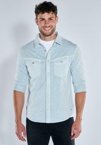 Light blue button-up shirt with two chest pockets, roll-up sleeves, and metallic buttons, worn over a white T-shirt. Smooth fabric texture.