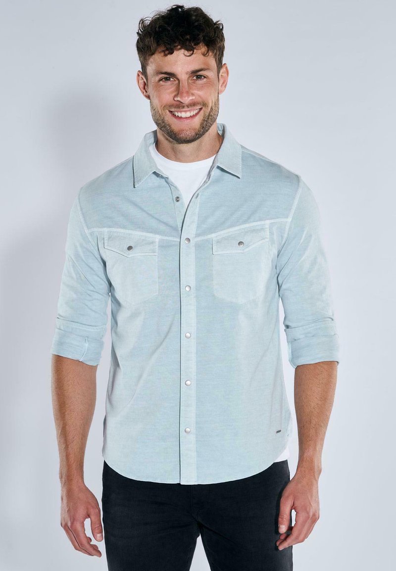 Light blue button-up shirt with two chest pockets, roll-up sleeves, and metallic buttons, worn over a white T-shirt. Smooth fabric texture.