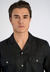 Chemise noire à manches courtes en tissu texturé. Dotée d'un col classique, de deux poches poitrine et de fermetures à boutons.