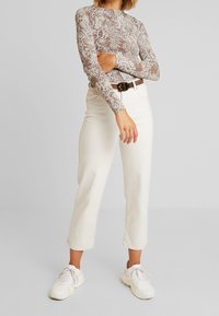 Langärmliges, enges Oberteil mit einem braunen und beigen Schlangenprint; kombiniert mit weißen High-Waist-Jeans und weißen Sneakers.