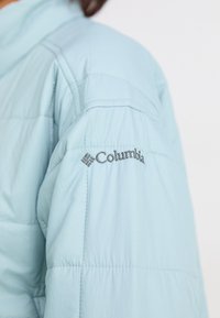 Šviesiai mėlynos paminkštintos striukės rankovė su siuvinėtu Columbia logotipu ant viršutinio rankovės, prieš baltą foną.
