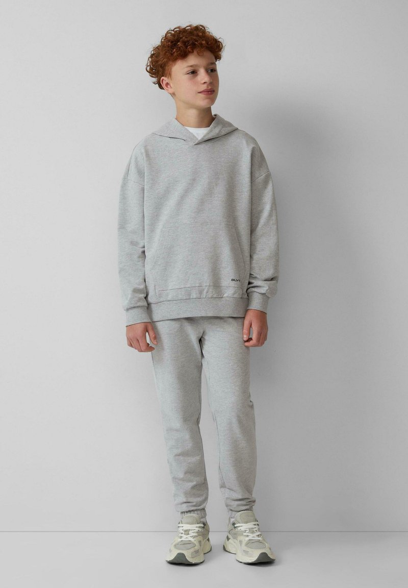 Grijze sweatshirt en sweatpants set van zachte stof. Met een capuchon en een voorzak. Model draagt lichte sneakers. Eenvoudig kleurenpalet.