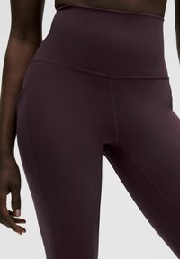 Nahaufnahme einer Person, die dunkelviolette High-Waist-Leggings mit sichtbaren Nähten trägt, während die entspannte Hand an der Seite ruht.