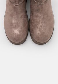 Bottes de cheville en daim marron avec une texture légèrement brillante, présentant des accents cousus et un design à bout arrondi, sur fond blanc.
