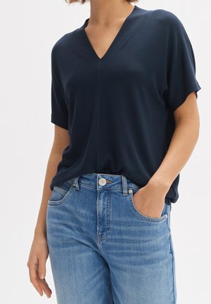 Femme portant un haut bleu marine à manches courtes avec un col en V, rentré dans un jean bleu clair, avec une main dans une poche, sur un fond blanc.