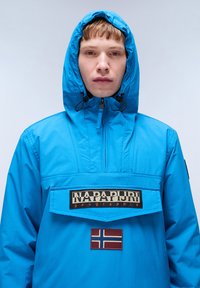 Giovane in giacca a zip di colore blu brillante con cappuccio, patch del logo Napapijri sul petto e patch della bandiera norvegese sotto.