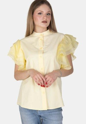 Giovane donna con lunghi capelli biondi indossa una camicetta giallo pallido con maniche a balze sovrapposte e jeans blu, in piedi davanti a uno sfondo bianco.