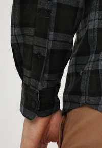 Camisa de franela a cuadros en negro y gris con mangas largas, que presenta botones negros y una tela texturizada, mostrada en la zona del puño y la muñeca.