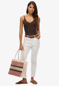 Bruine gebreide tanktop, witte strakke jeans, bruine riem en bruine sandalen. Houdt een gestreepte totebag met het "SUPERDRY" logo in het geel.