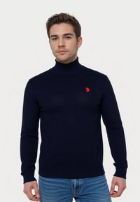 Joven con un suéter de cuello alto azul marino con un pequeño logo rojo en el pecho y jeans de mezclilla claros, de pie frente a un fondo sencillo.