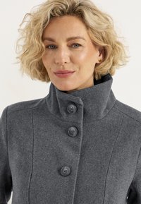 Grau wollener Mantel mit hohem Kragen, drei großen schwarzen Knöpfen und einer strukturierten Stoffoberfläche. Taillierte Passform mit vertikalen Nahtdetails.