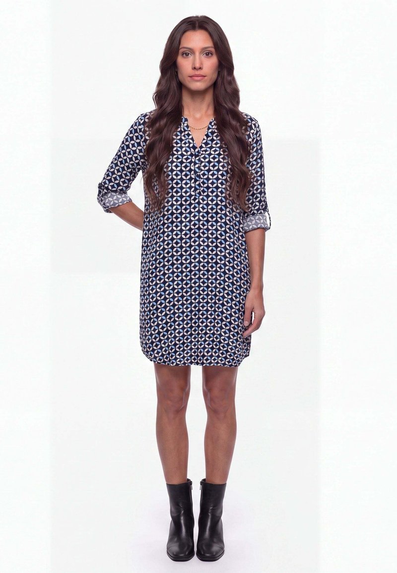 Femme aux longs cheveux foncés portant une robe bleue à motifs longueur genoux et des bottines noires, debout devant un fond blanc.