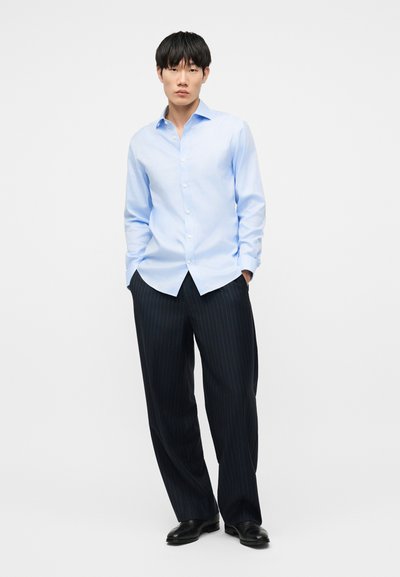 Άνδρας που στέκεται με τα χέρια στις τσέπες, φοράει ανοιχτό μπλε πουκάμισο με κουμπιά, μαύρο παντελόνι με ρίγες και μαύρα παπούτσια σε λευκό φόντο.