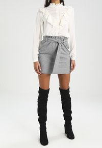 Bílá blůza s volánkovým límcem v kombinaci se šedou kostkovanou mini sukni s ozdobným páskem, doplněná černými kozačkami nad kolena.