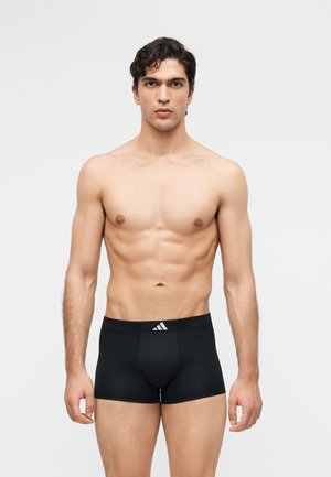 Noor mees lühikeste lokkis juustega, mustade Adidas bokseripükstega, seisab tühja heleda tausta ees.